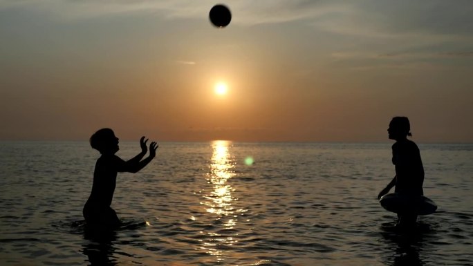 日落时分，一男一女两个少年站在海边打排球的剪影。快乐的弟弟和妹妹用手掌把球推向对方。水排球