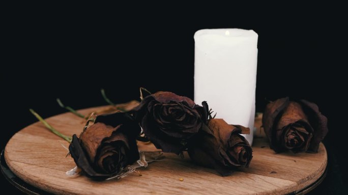燃烧的白色蜡烛和四朵枯萎的干玫瑰在黑色背景上旋转