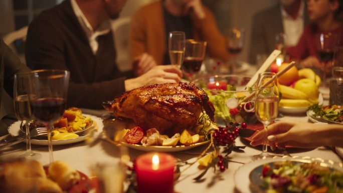 节日感恩节晚餐或圣诞庆祝家庭餐。人们坐在餐桌后面，享用火鸡大餐、烤土豆、配菜和饮料。美食的特写镜头