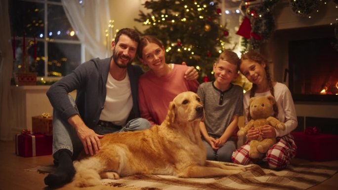 一个快乐的年轻家庭在镜头前摆姿势，微笑着互相拥抱的圣诞肖像。丈夫、妻子、女儿、儿子和一只宠物金毛猎犬