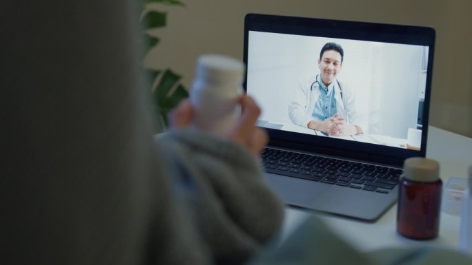 亚洲年轻女孩患者的特写，在笔记本电脑上咨询医生视频通话，在应用程序上进行药物剂量说明，夜间在家远程医