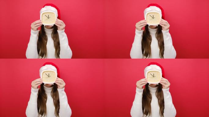 可爱的年轻女子穿着温暖的毛衣，手里拿着圣诞老人的帽子，脸上戴着时钟显示5点到12点，在淡红色的工作室