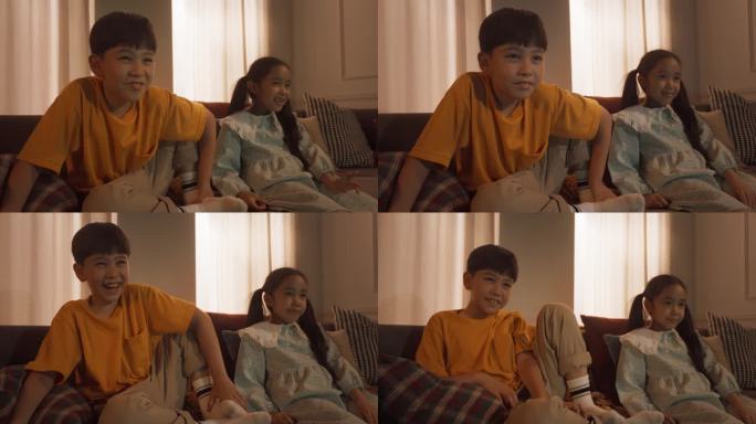 可爱的小兄弟姐妹在舒适的客厅沙发上看电视，开心地笑着。韩国兄弟姐妹晚上在家享受儿童电视节目或动画片。
