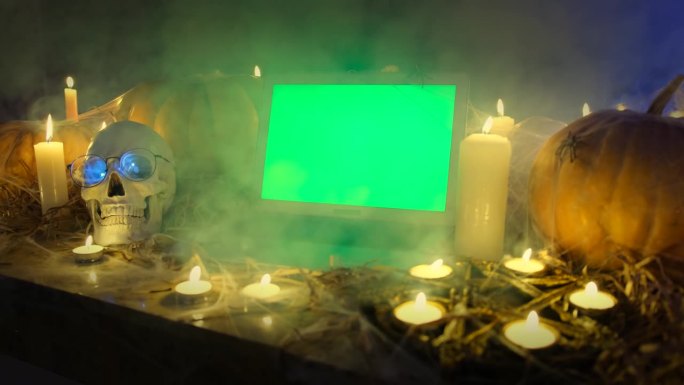 笔记本电脑与绿色屏幕在雾和万圣节橙色南瓜，蜡烛，头骨，五角星蛛网在干草在黑暗的精神之夜