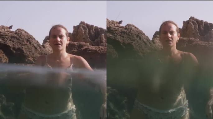 垂直视频。一名年轻女子站在岩石上透明的海水中，相机放置在水边，捕捉水面上和水下的画面，放大了水下场景