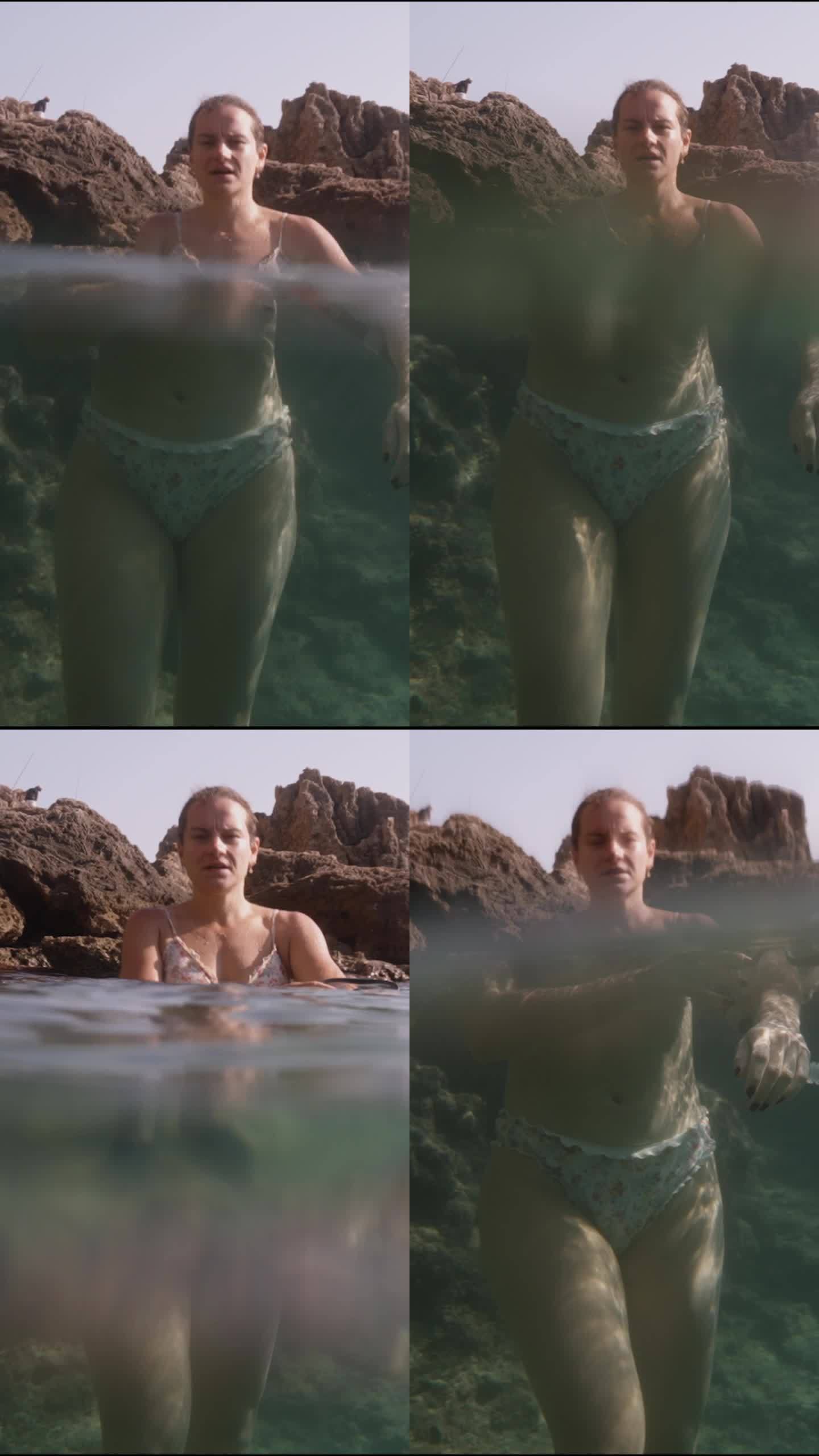 垂直视频。一名年轻女子站在岩石上透明的海水中，相机放置在水边，捕捉水面上和水下的画面，放大了水下场景