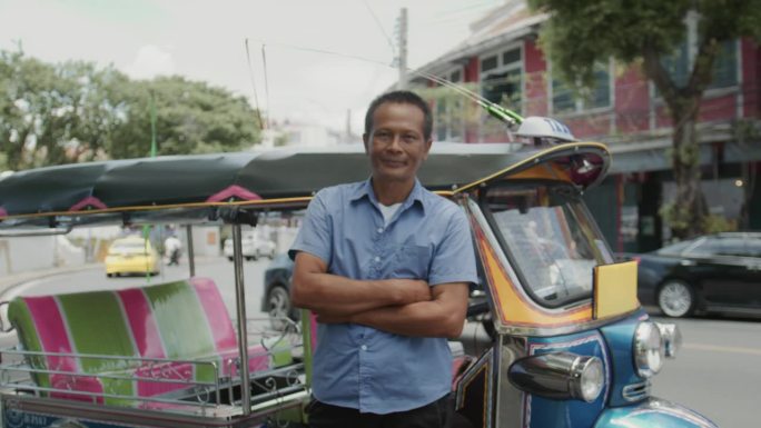 一名亚洲男子嘟嘟车司机站在那里，双臂交叉，微笑着看着镜头