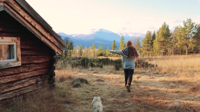 穿着羊毛衫的快乐女人带着狗在木屋边跑步，在挪威的群山中欣赏日落