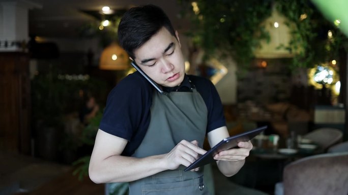 聪明的服务员穿着朴素的制服，用智能手机和平板电脑接单。穿着深色围裙的成功男性餐厅老板通过互联网向外卖
