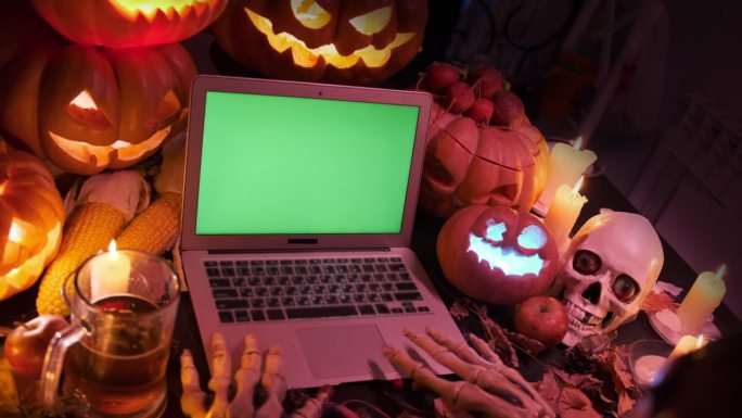 令人毛骨悚然的骷髅手使用着绿色屏幕的笔记本电脑，躺在黑暗中闪烁的万圣节雕刻南瓜灯的装饰品旁边