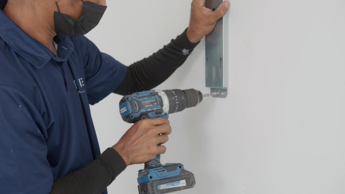 一名亚洲男子用无绳钻把螺丝拧到墙上，在客户家里安装电力系统