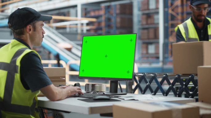 白人男性配货助理在配货设施使用绿屏Chromakey台式电脑。多民族男性仓库工人在自动传送带上装载纸