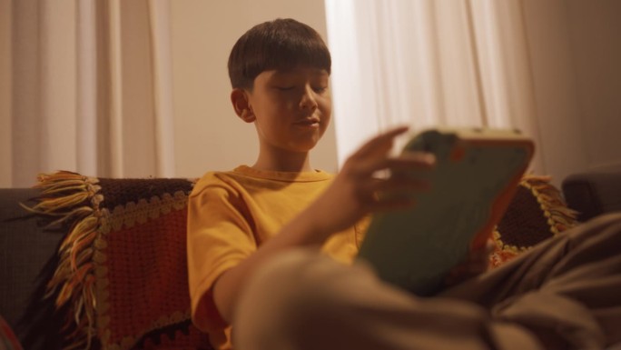 一个韩国小男孩坐在客厅的沙发上，用平板电脑玩电子游戏的低角度肖像。活跃的男孩使用互动媒体学习