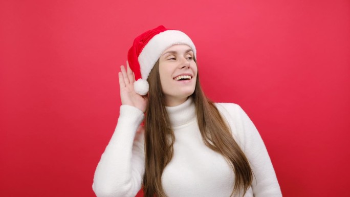 好奇的爱管闲事的年轻女子穿着温暖的白色毛衣，戴着圣诞老人帽，试着听你偷听，在工作室里淡红色的背景下独