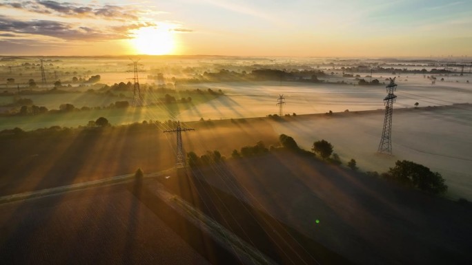 空中拍摄的工业公用事业电线和塔结构在雾蒙蒙的农业树篱景观的早晨日出。多条电力线和电线塔横跨农田。