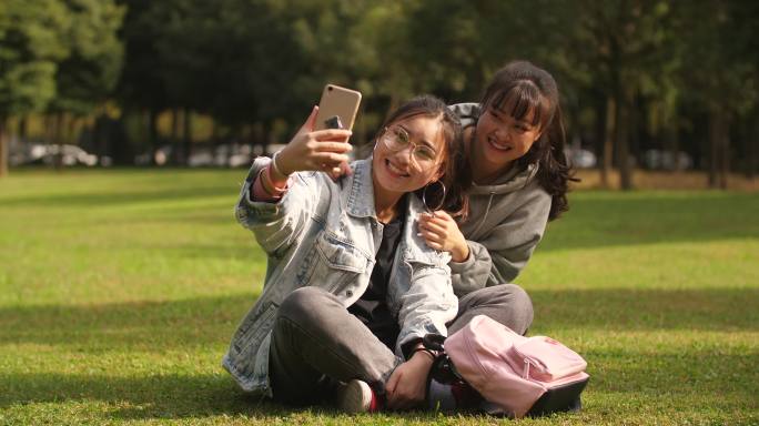 两位开心的女大学生坐在校园草坪上手机自拍