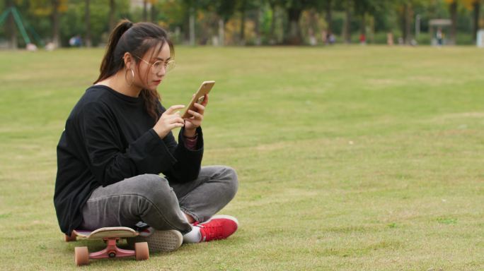 大学校园草地上女大学生坐在滑板上玩手机