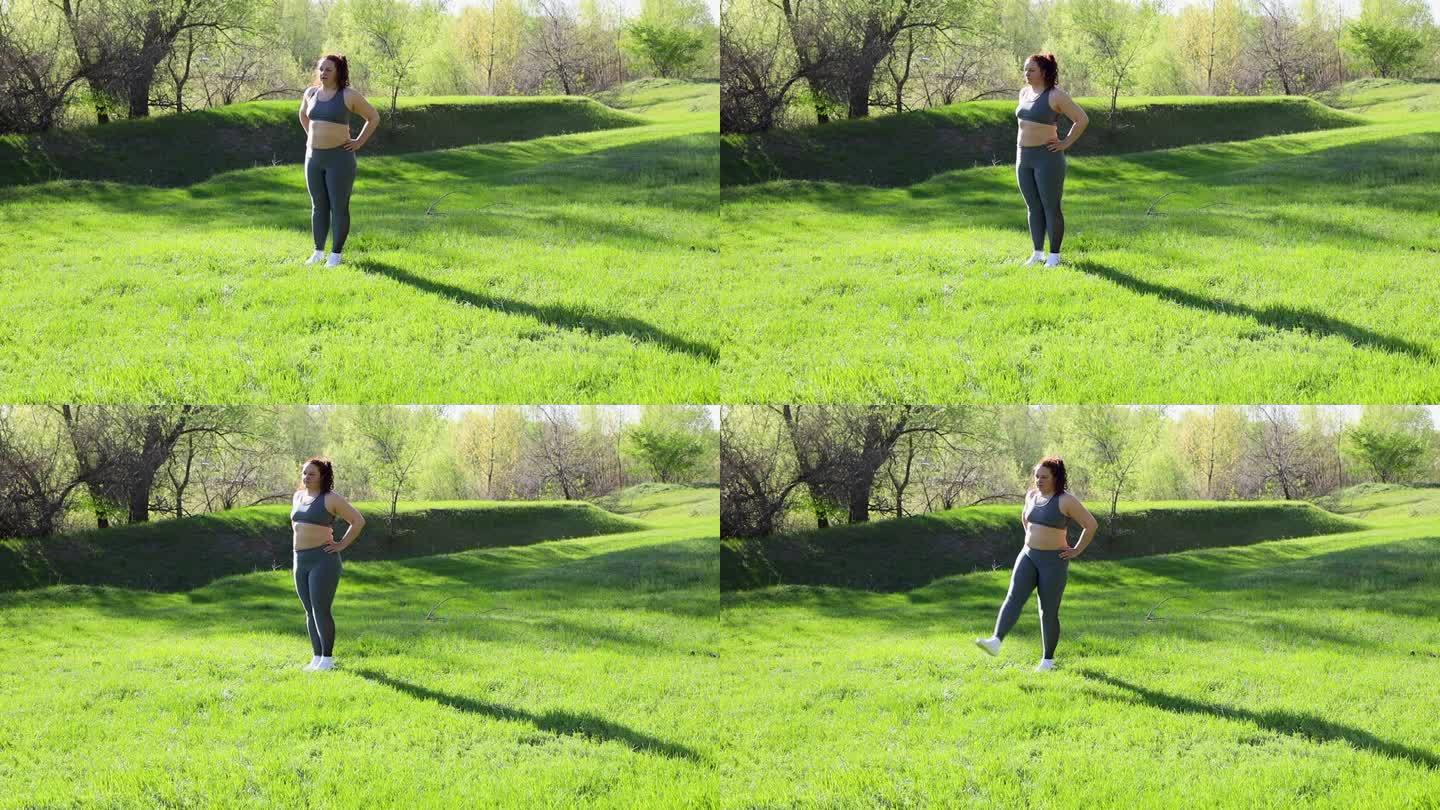 在阳光明媚的日子里，穿着健身服的年轻超重女子站在公园的绿色草坪上，双脚向前摆动，背景是大自然。健康的