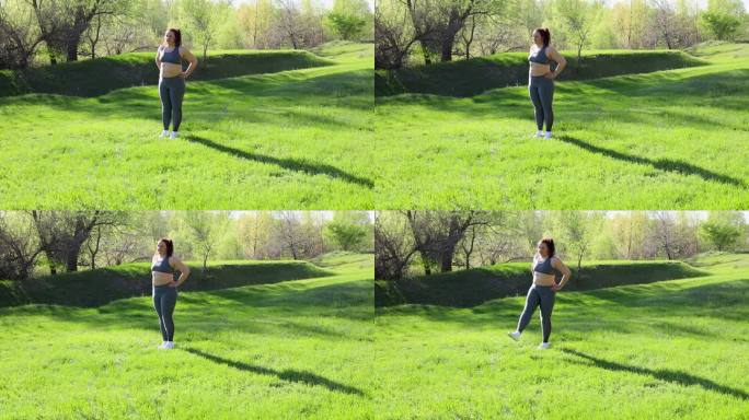 在阳光明媚的日子里，穿着健身服的年轻超重女子站在公园的绿色草坪上，双脚向前摆动，背景是大自然。健康的