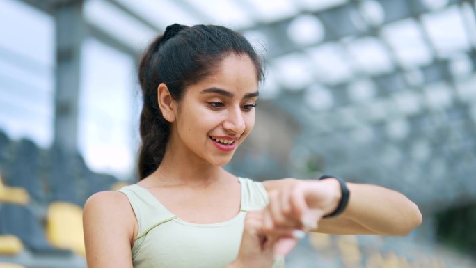 快乐的年轻女运动员在城市体育场看智能手表。女运动员使用检查健身追踪器