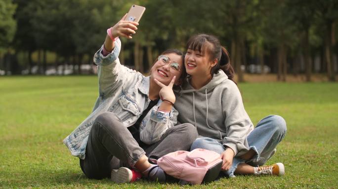 两位开心的女大学生坐在校园草坪上手机自拍