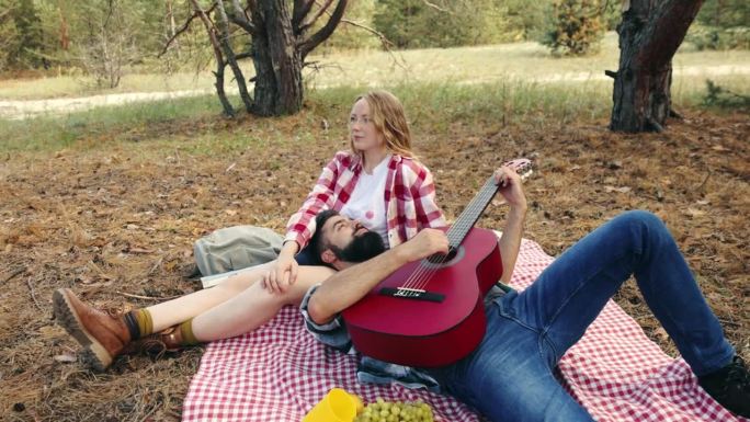 热恋的年轻夫妇，男人和女人在温暖的阳光下在森林里野餐。一个男人为他漂亮的女朋友弹吉他