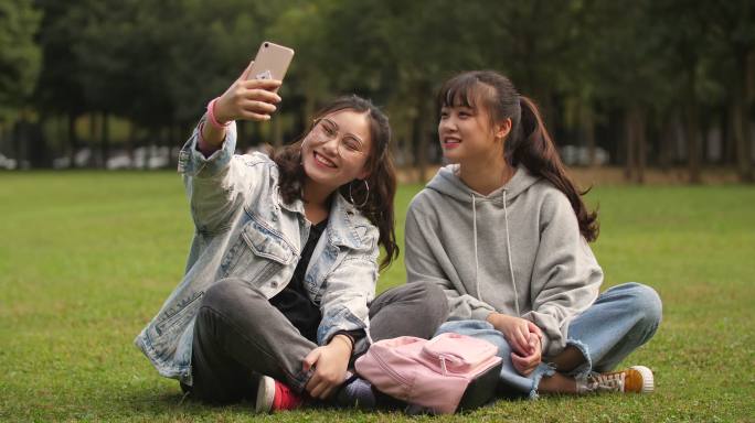 两位开心的女大学生坐在校园草坪上手机自拍