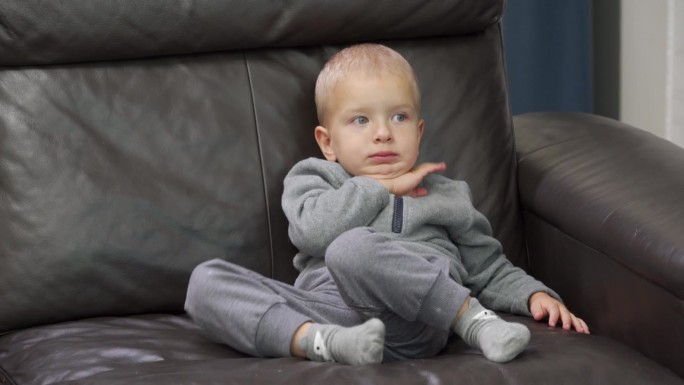 蹒跚学步的孩子小男孩坐在家里的皮沙发上看电视