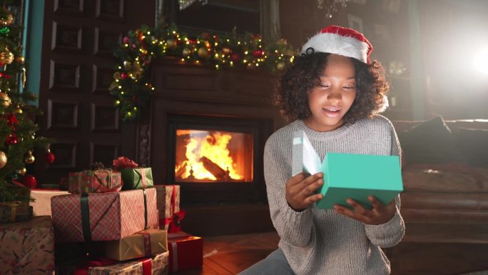 圣诞快乐。一位非裔美国妇女在圣诞树旁打开礼品盒。女孩在客厅用圣诞树和壁炉打开礼盒带着惊喜的脸，平安夜