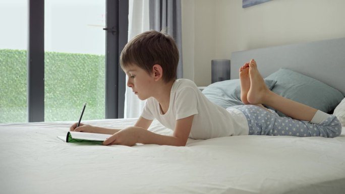 小男孩穿着睡衣躺在大窗前的床上，在笔记本上做作业。教育的概念，儿童发展和远程教育在家里