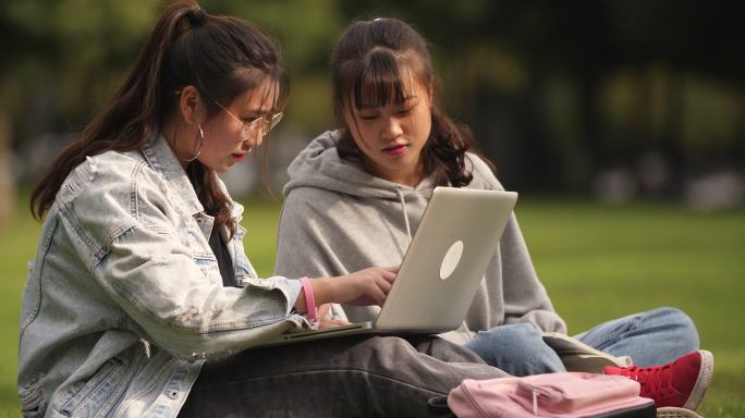 两位女大学生坐在校园草地上用电脑讨论学习