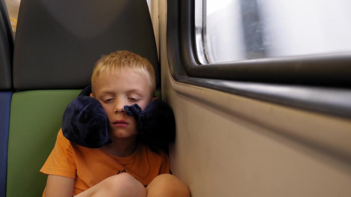 一个抱着颈枕的小男孩坐在火车上靠窗的地方睡觉。