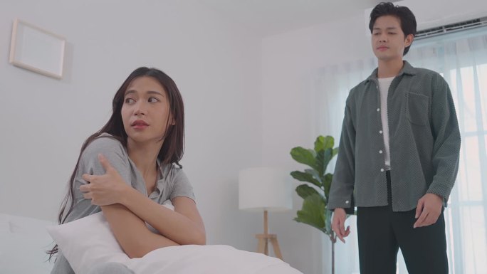 亚洲年轻夫妇坐在床上痛苦的争吵后打架。新婚男女因在卧室里吵架而伤心欲绝。紧张的亚洲女人坐在床上哭