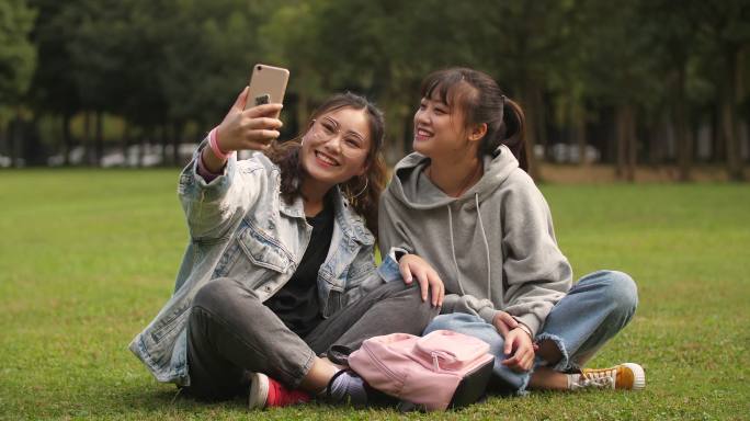 两位开心的女大学生坐在校园草坪上手机自拍