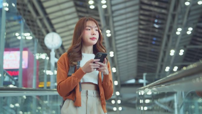 亚洲女游客背着背包，站在机场候机楼的移动走道上使用手机，旅游旅行的概念。