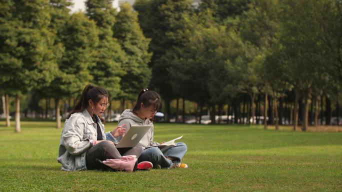 两位女大学生坐在校园草地上用电脑讨论学习