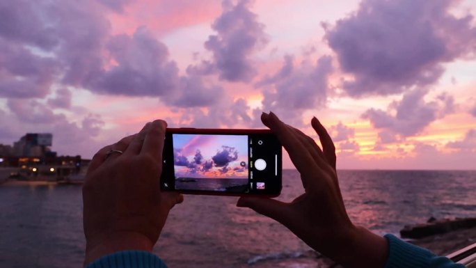女人手里拿着智能手机，拍着海上的夕阳