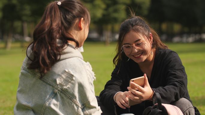 两位女大学生坐在校园草地上看手机聊天