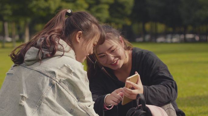 两位女大学生坐在校园草地上看手机聊天