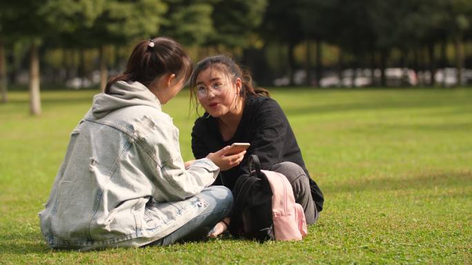 两位女大学生坐在校园草地上看手机聊天