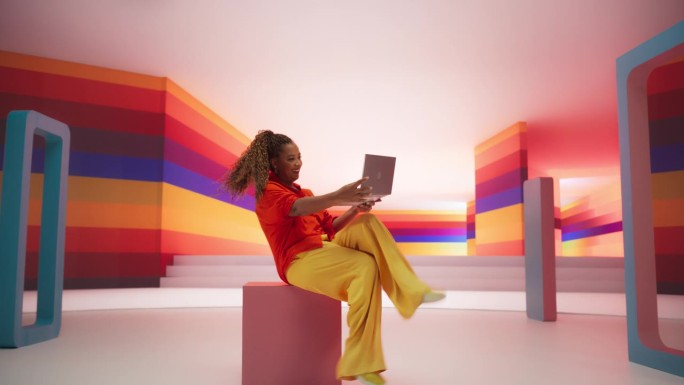 穿着鲜艳衣服的黑人女性坐在几何抽象环境中使用笔记本电脑。富有创造力的年轻女性在色彩缤纷的工作室里快乐