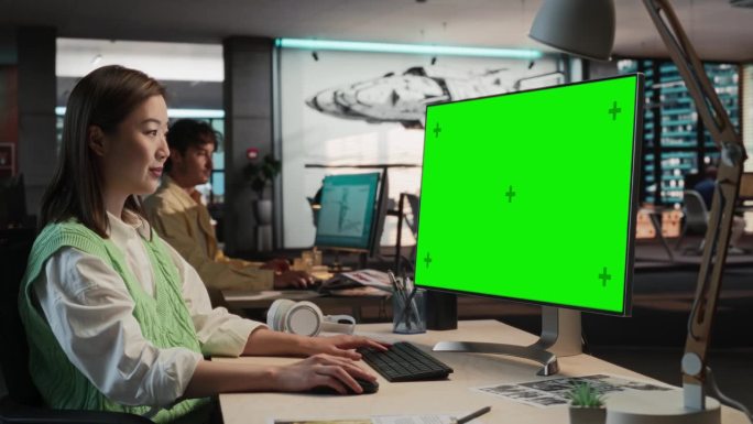 肖像的亚洲妇女坐在她的办公桌上使用台式电脑与模拟绿屏铬。在游戏开发工作室工作的女性概念美工，创造沉浸