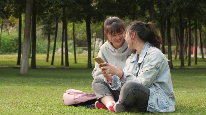 两位女大学生坐在校园草地上看手机学习交谈