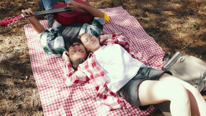 热恋的年轻夫妇，男人和女人在温暖的阳光下在森林里野餐。躺在毯子上，男人在弹吉他