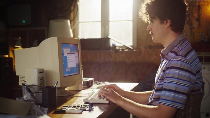 白人男性工程专业学生在复古车库用旧台式电脑写学术论文的侧视图。未来工程师聚焦九十年代科技大学创新项目