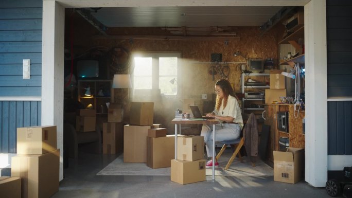 西班牙裔女商人在车库使用笔记本电脑。女小企业主填写网上客户的订单。产品在纸板箱准备运输。在家工作的企
