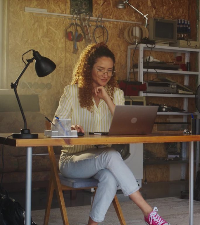 垂直屏幕:西班牙裔女程序员在家里车库的笔记本电脑上编程。年轻女性致力于软件服务创业。创办科技公司的数