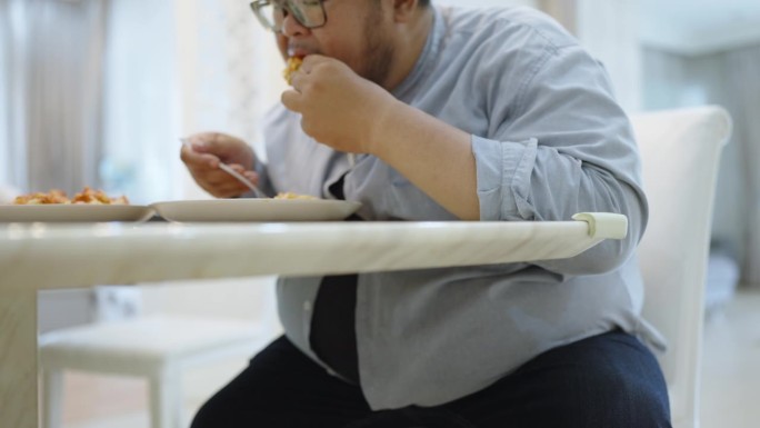 一个身材魁梧的男人坐在餐桌旁，用叉子狼吞虎咽地吃着意大利肉酱面，用手咬着炸鸡翅。