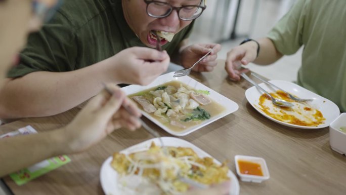 从肩膀上看，两名男子正在吃面条和脆口贻贝煎蛋卷，而另一名男子在小吃摊上吃午饭时几乎吃光了他的米饭。