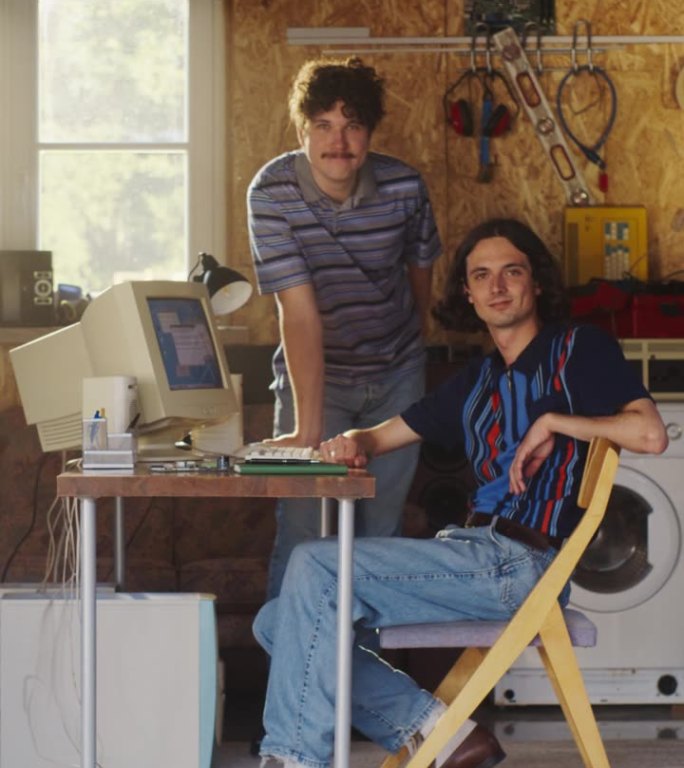 垂直屏幕:两个白人男性科技创业公司创始人在复古车库使用旧台式电脑，看着镜头微笑。程序员和用户体验设计